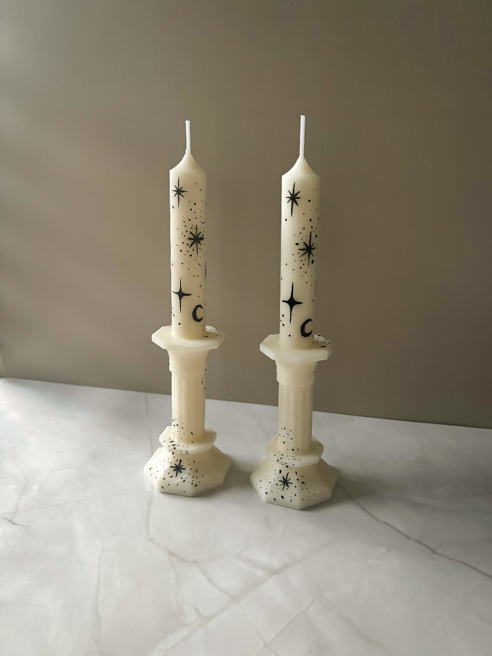 Two white taper candles with hand painted black star patterns on a marble surface against a beige wall.